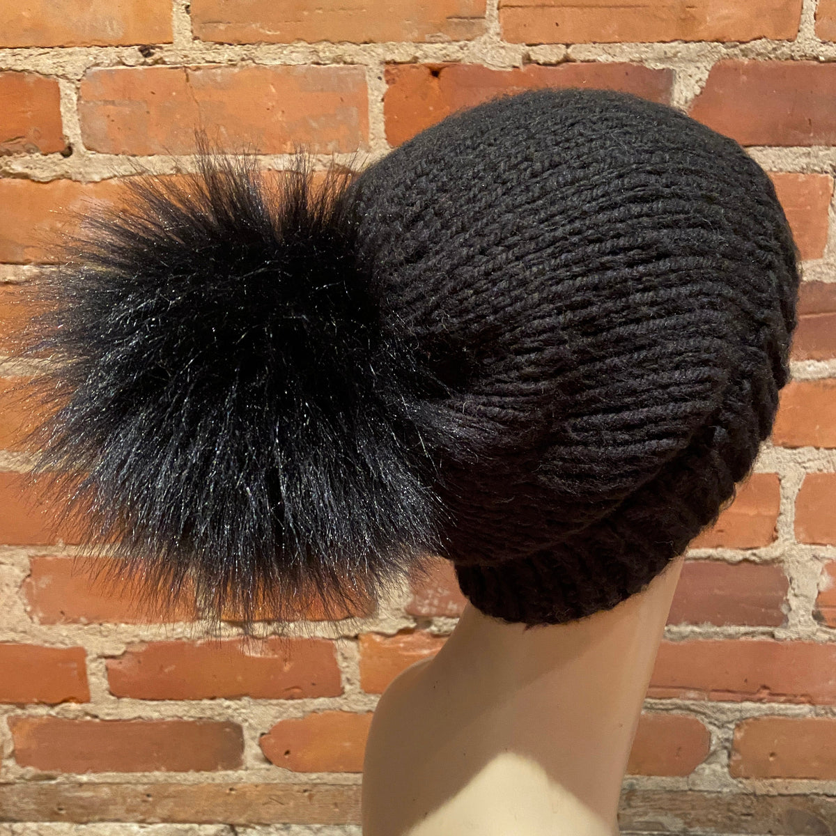 A black ribbed knit beanie hat with a large fluffy black faux-fur pom-pom, displayed on a mannequin head against a red brick wall background.