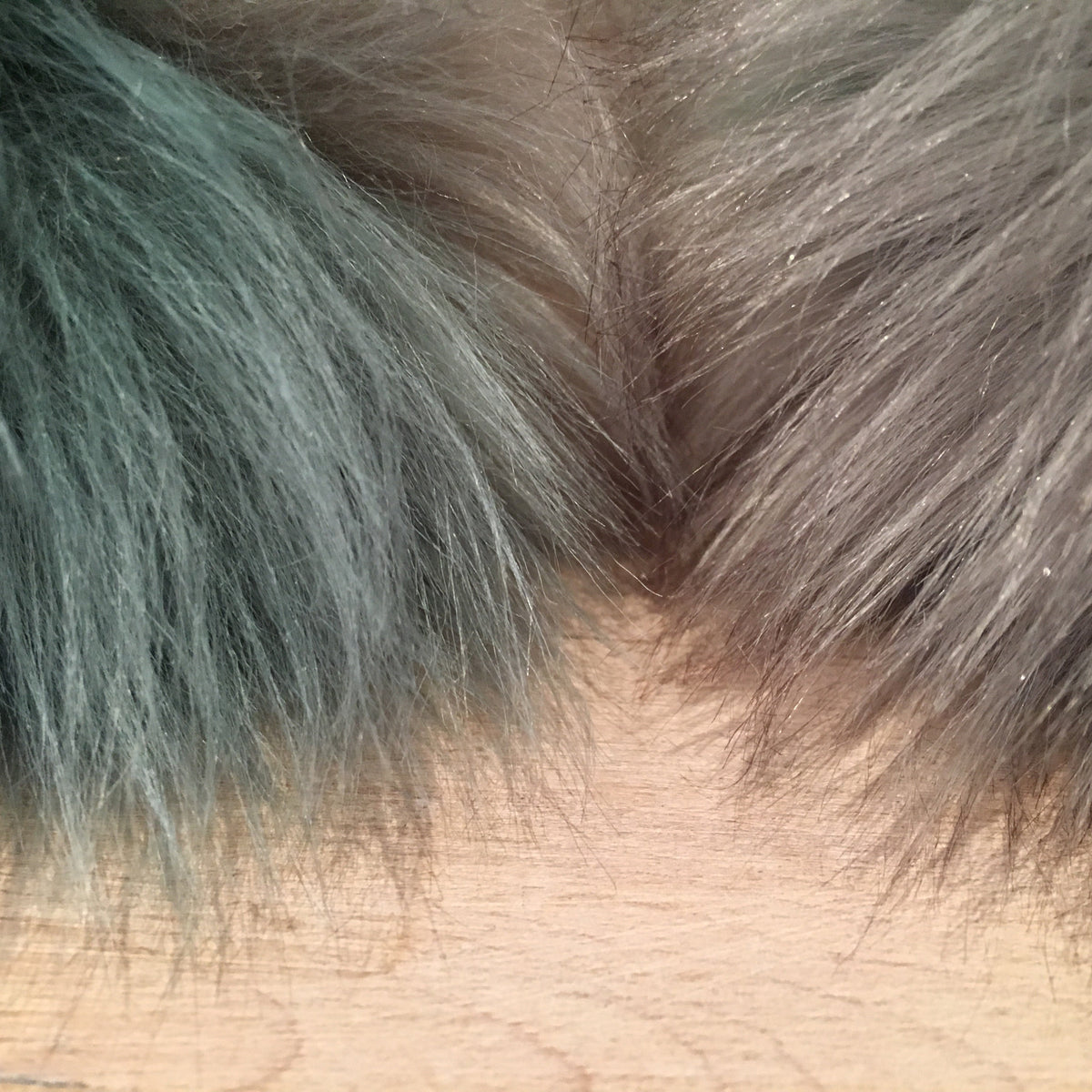 Close-up of two faux fur fabric swatches, one in muted teal-blue and one in taupe-brown, meeting at a V-shape on a light wood surface.