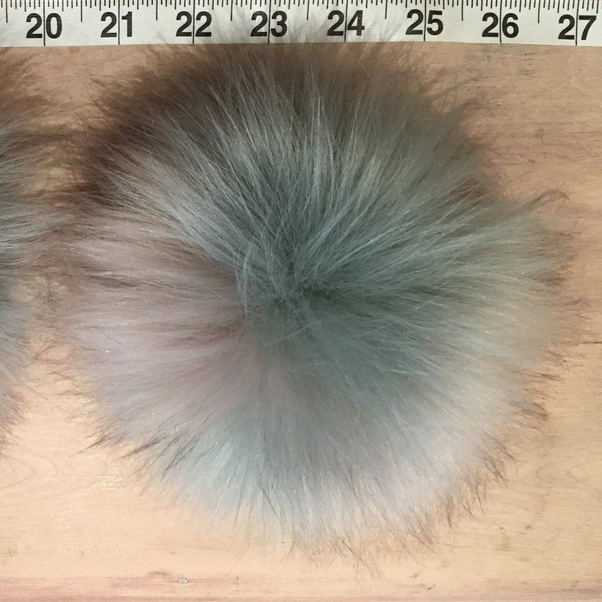 Two fluffy faux-fur pom-poms in a mottled gray and white colorway, displayed side-by-side on a wooden surface with a measuring tape visible at the top for scale.