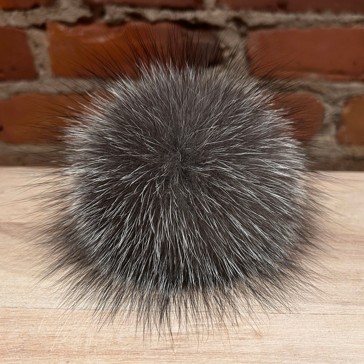 A round, fluffy fox -fur pom-pom in a gray and white mottled pattern, resting on a light wood surface with a blurred red brick wall background.