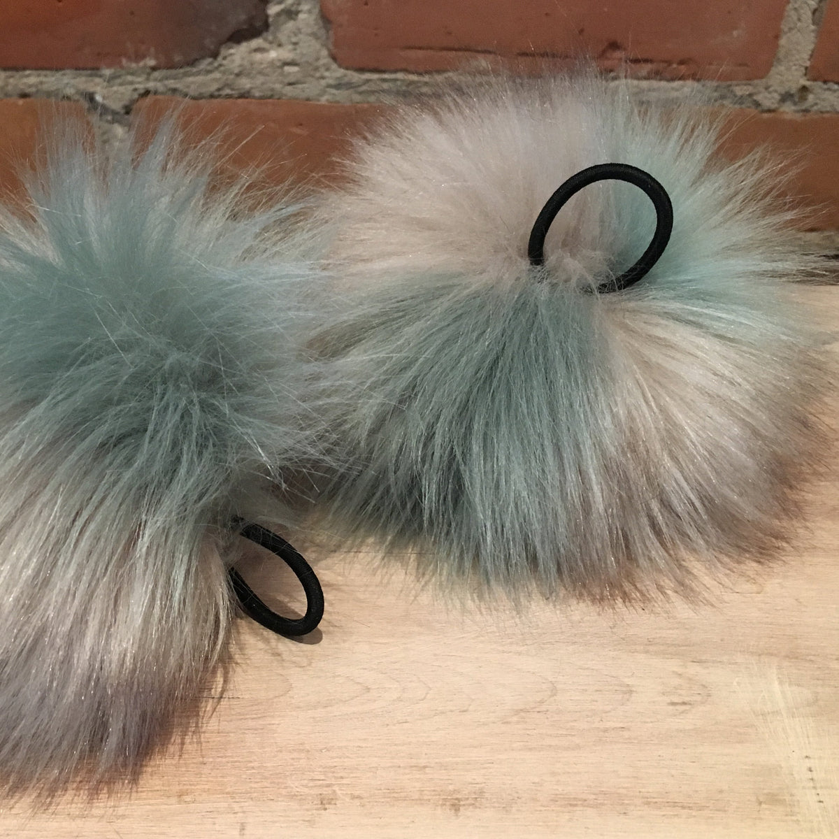 Two fluffy faux-fur pom-pom accessories in a pale sage green-gray color, each attached to a black elastic band, displayed on a wooden surface against a brick wall background.
