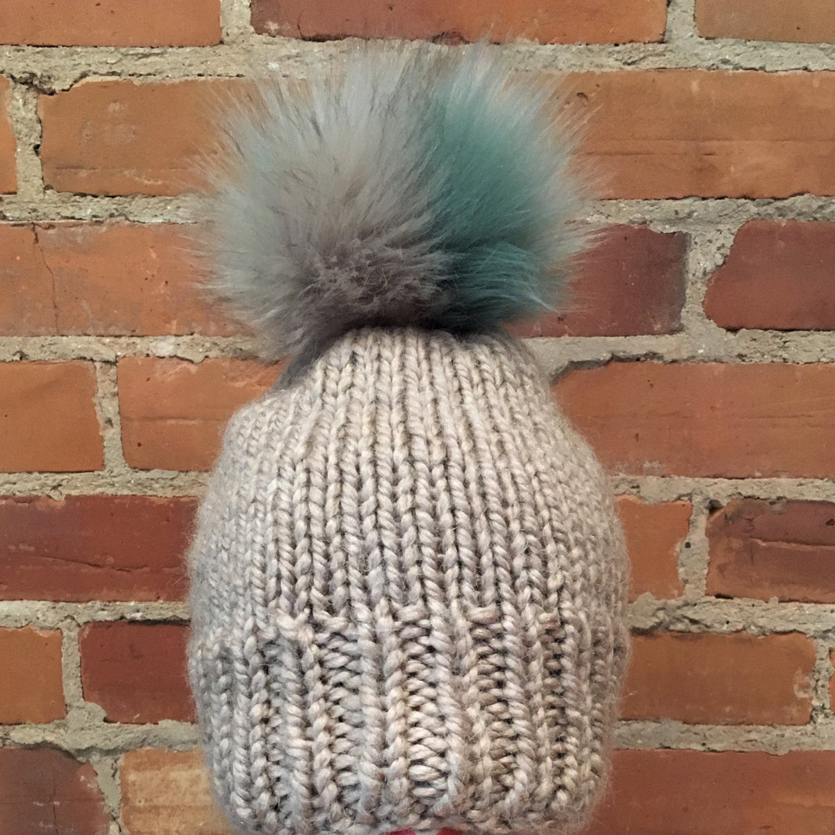 A chunky knit beanie in light gray with a large fluffy pom-pom in gray and mint green, displayed on a white mannequin head against a red brick wall background.