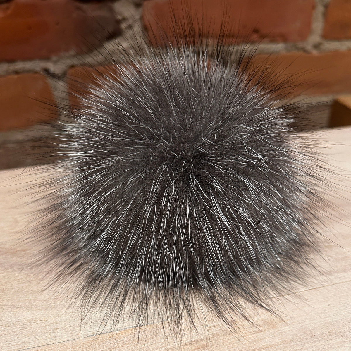 A round, fluffy fox -fur pom-pom in a dark gray and silver-gray color, resting on a light wood surface with a brick wall background.