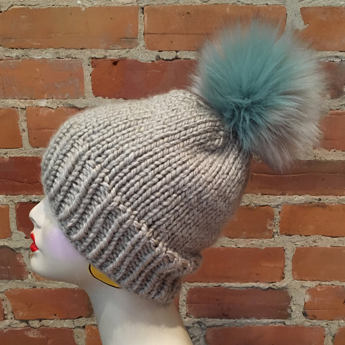A light gray ribbed knit beanie with a large fluffy teal and gray pom-pom, displayed on a mannequin head against a red brick wall background.