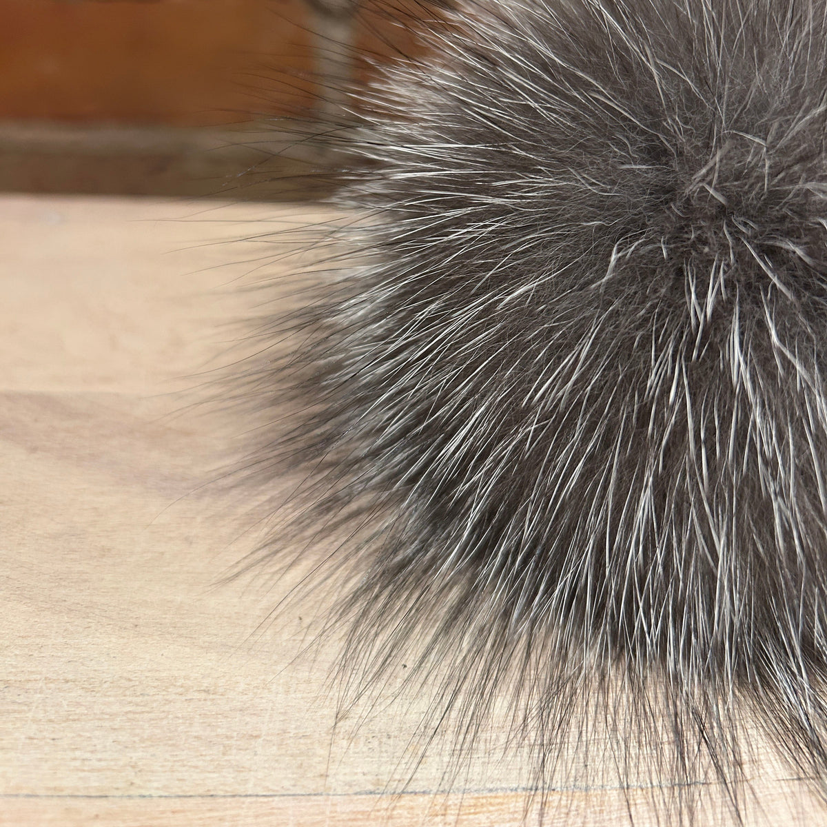 A close-up of a fluffy gray and white fox fur pom-pom resting on a light wood surface with a blurred brick wall background.
