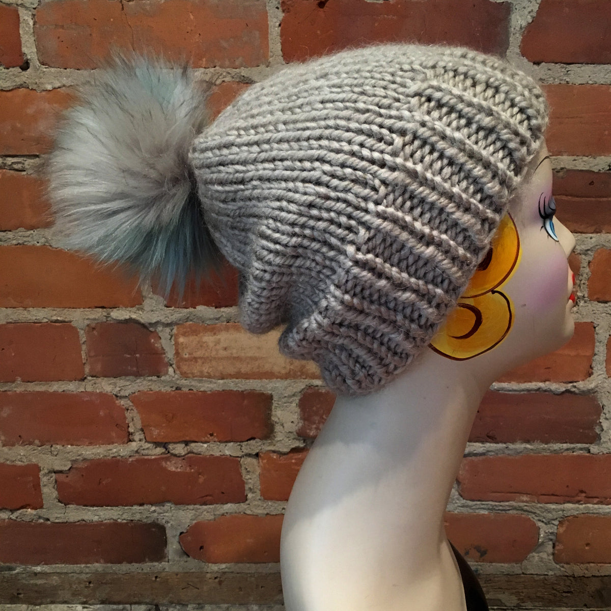 A chunky knit slouchy beanie hat in a light gray color with a large fluffy pom-pom in a mix of gray, white, and light blue, displayed on a mannequin head against a red brick wall background.