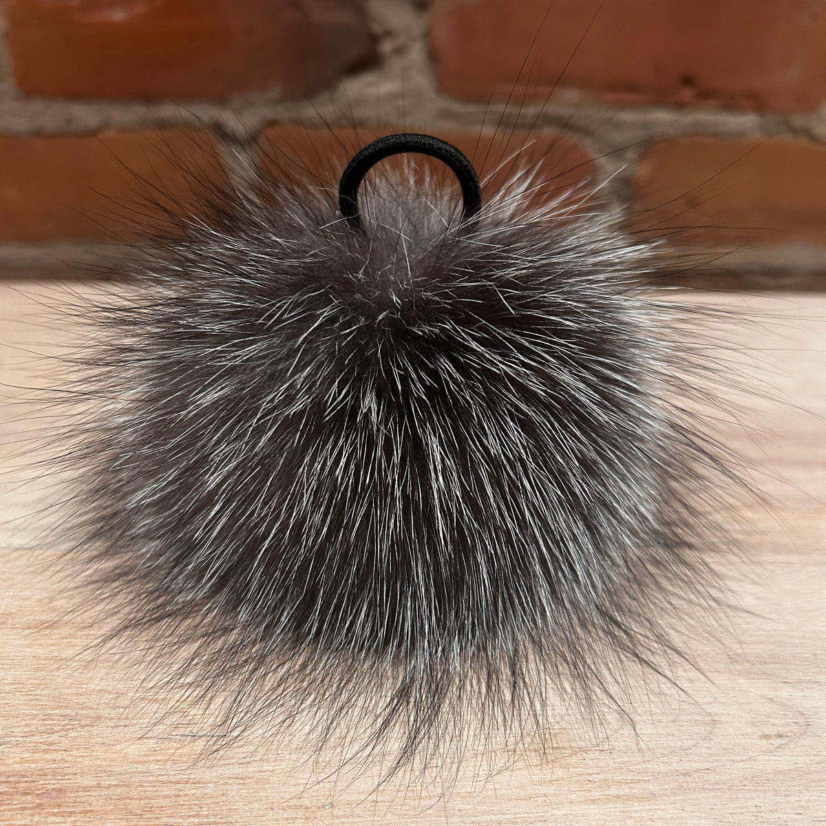 A fluffy fox -fur pom-pom keychain or bag charm in dark gray and black, attached to a black elastic loop, resting on a light wood surface with a brick wall background.