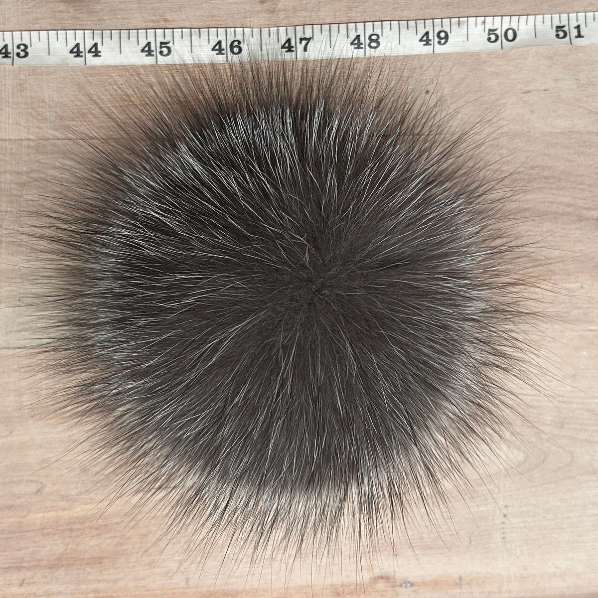 A round, fluffy fox fur pom-pom in dark gray and black, shown on a light wood background with a measuring tape ruler at the top for scale.