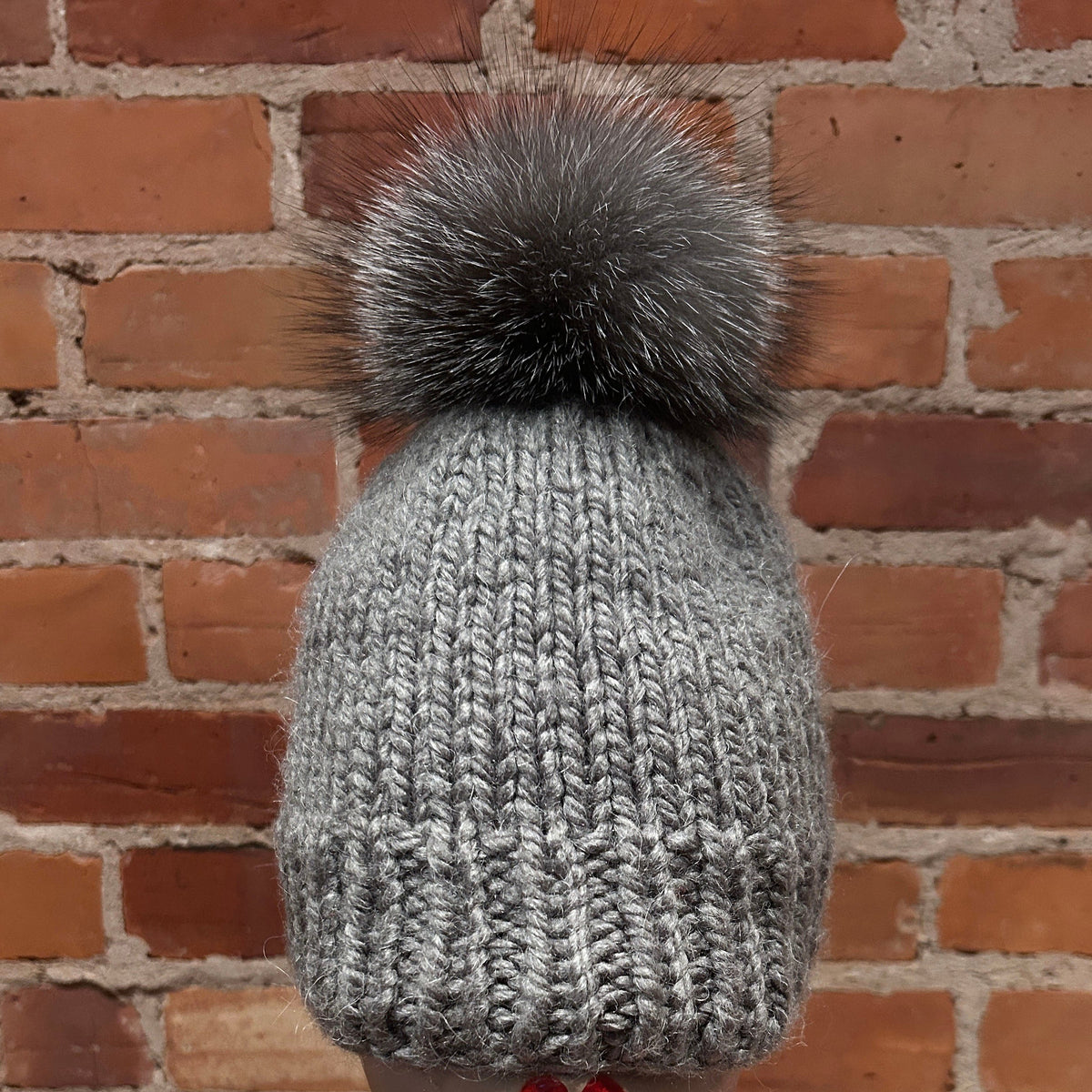 A gray ribbed knit beanie with a large gray fox fur pom-pom, displayed on a mannequin head against a red-brown brick wall background.
