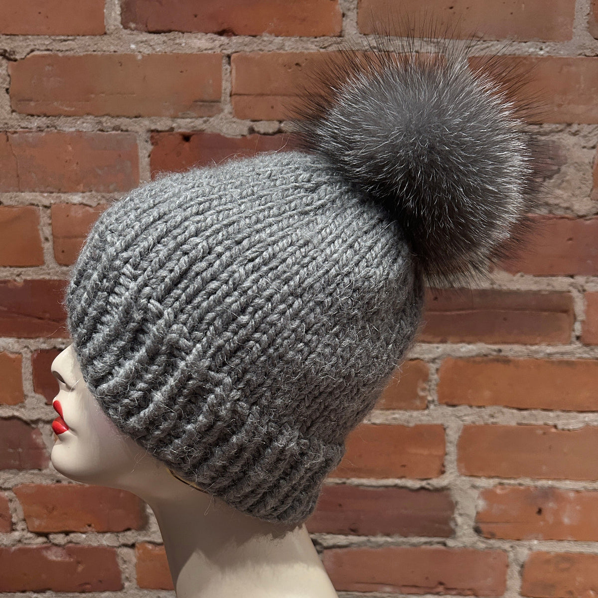 A gray ribbed knit beanie with a large fluffy gray pom-pom, displayed on a mannequin head in profile against a red brick wall background.