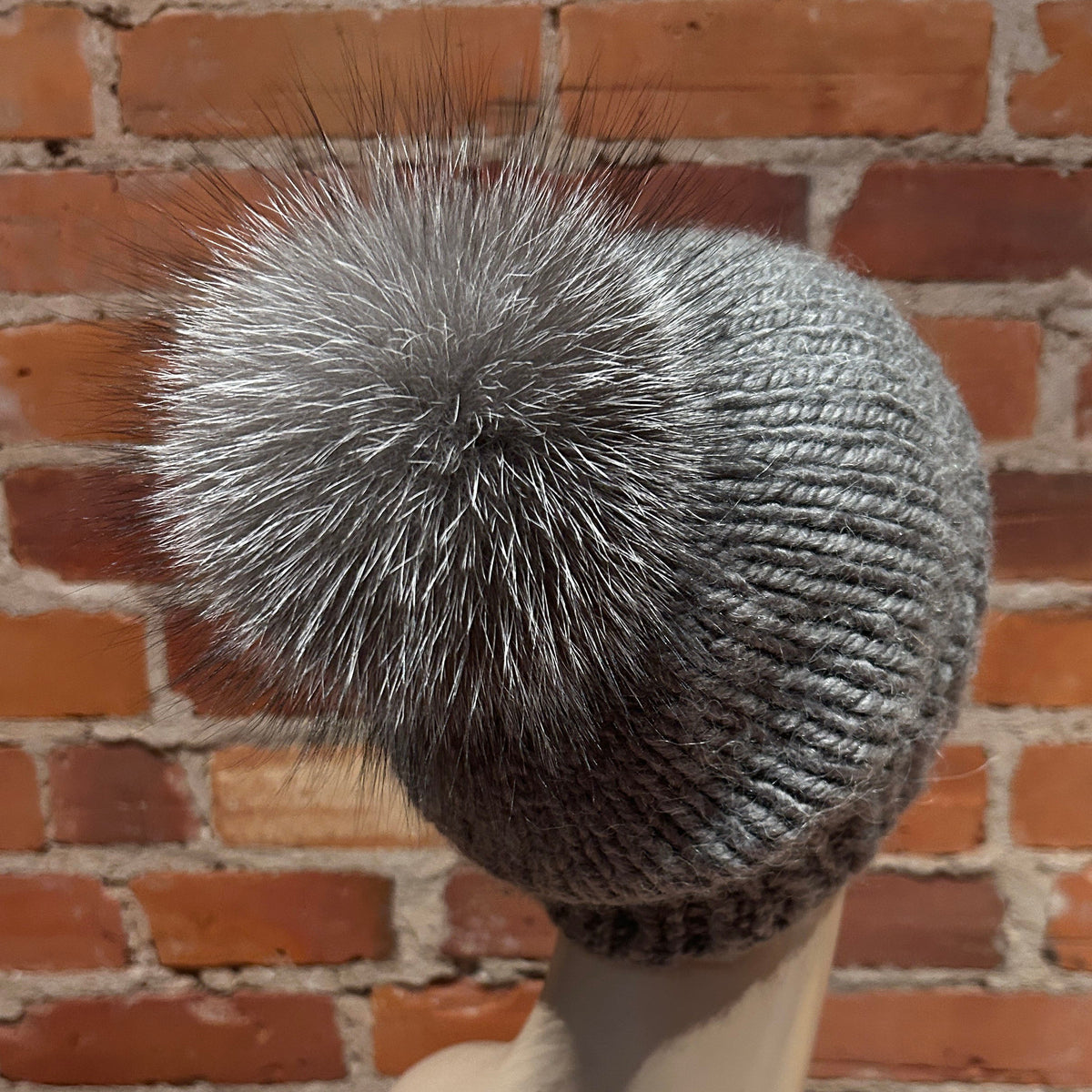 A gray ribbed knit beanie with a large fluffy silver-gray fox fur pom-pom, displayed on a mannequin head against a red brick wall background.
