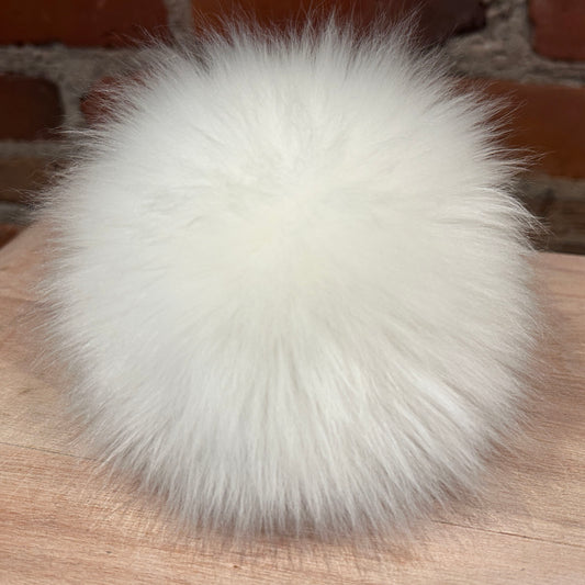 A fluffy white lamb fur pom-pom ball resting on a light wood surface with a brick wall background.