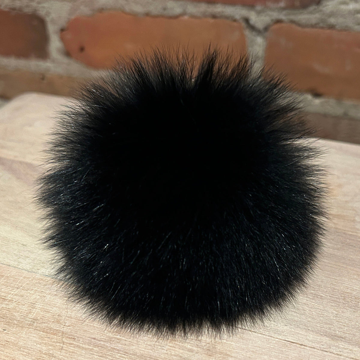 A round, fluffy black fox fur pom-pom resting on a light-colored wooden surface with a brick wall in the background.