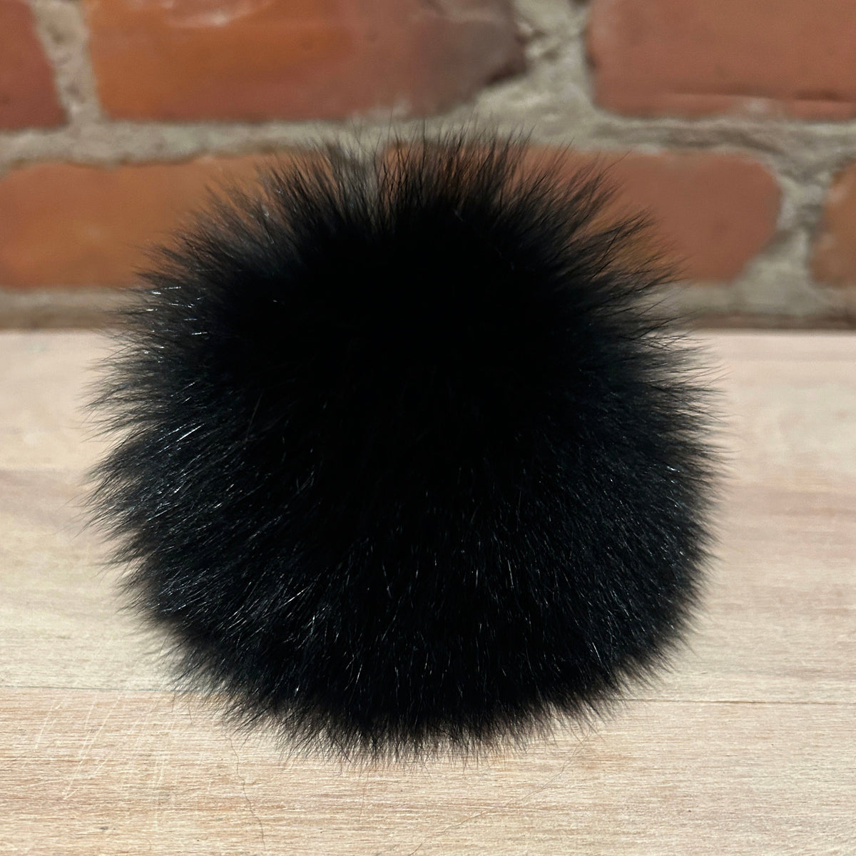 A round, fluffy black fox fur pom-pom accessory resting on a light wood surface with a brick wall background.