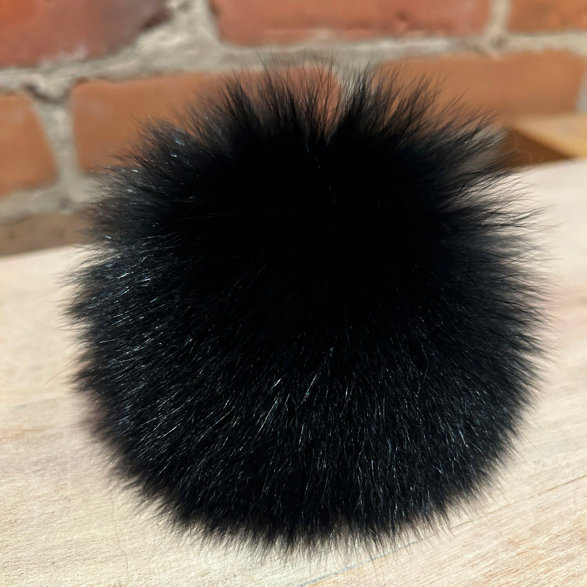 A fluffy black fox fur pom-pom accessory resting on a light wood surface with a brick wall background.