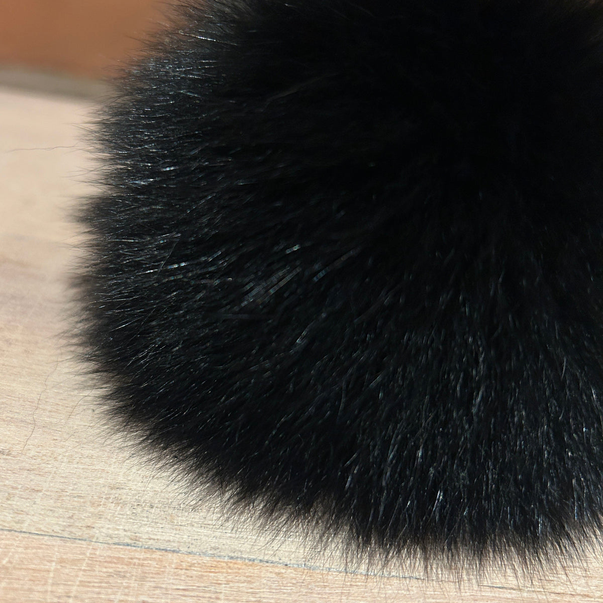 A close-up of a fluffy black fox fur pom-pom resting on a light wood surface with a blurred brick wall in the background.