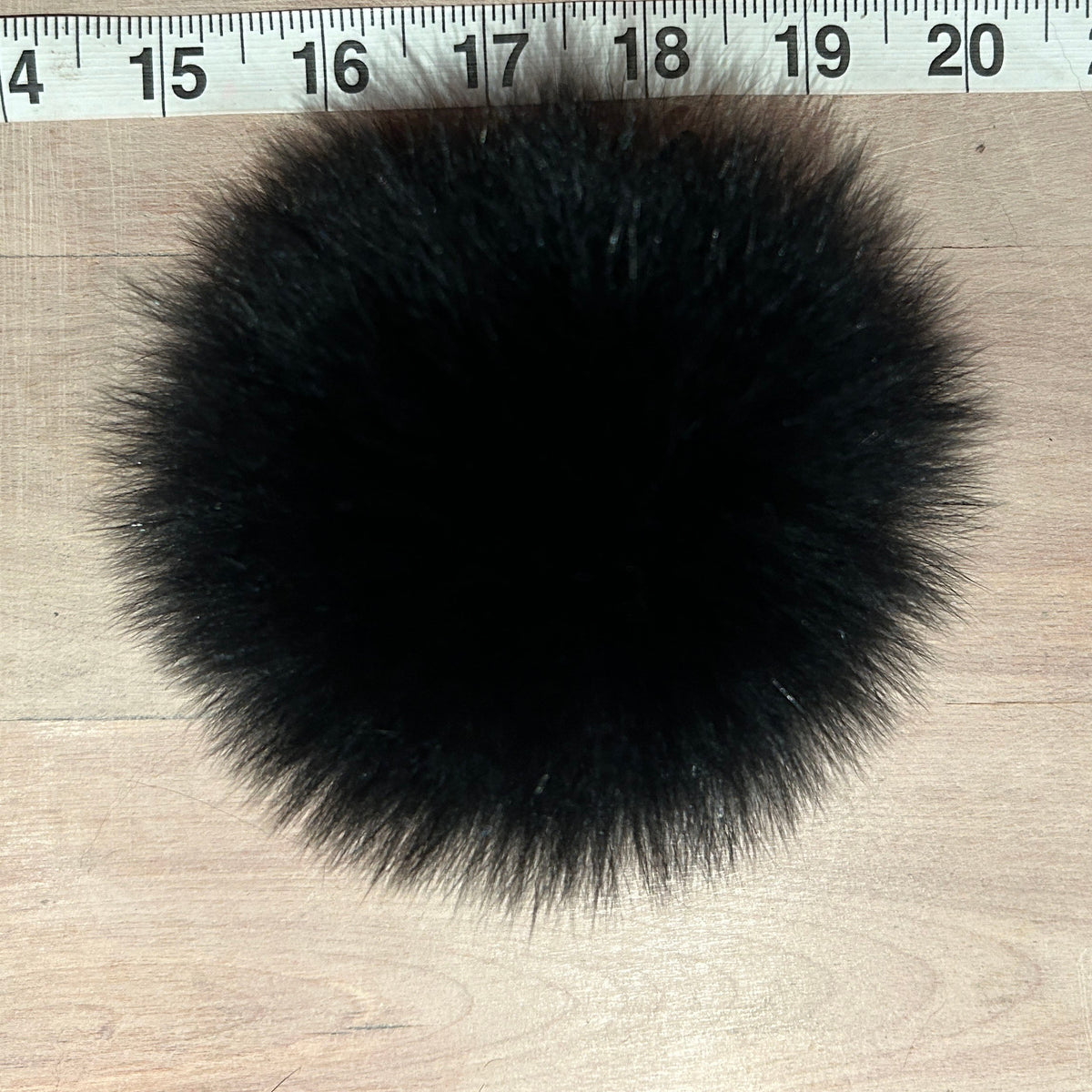 A round black fox fur pom-pom accessory shown on a light wood surface next to a measuring tape ruler for scale.