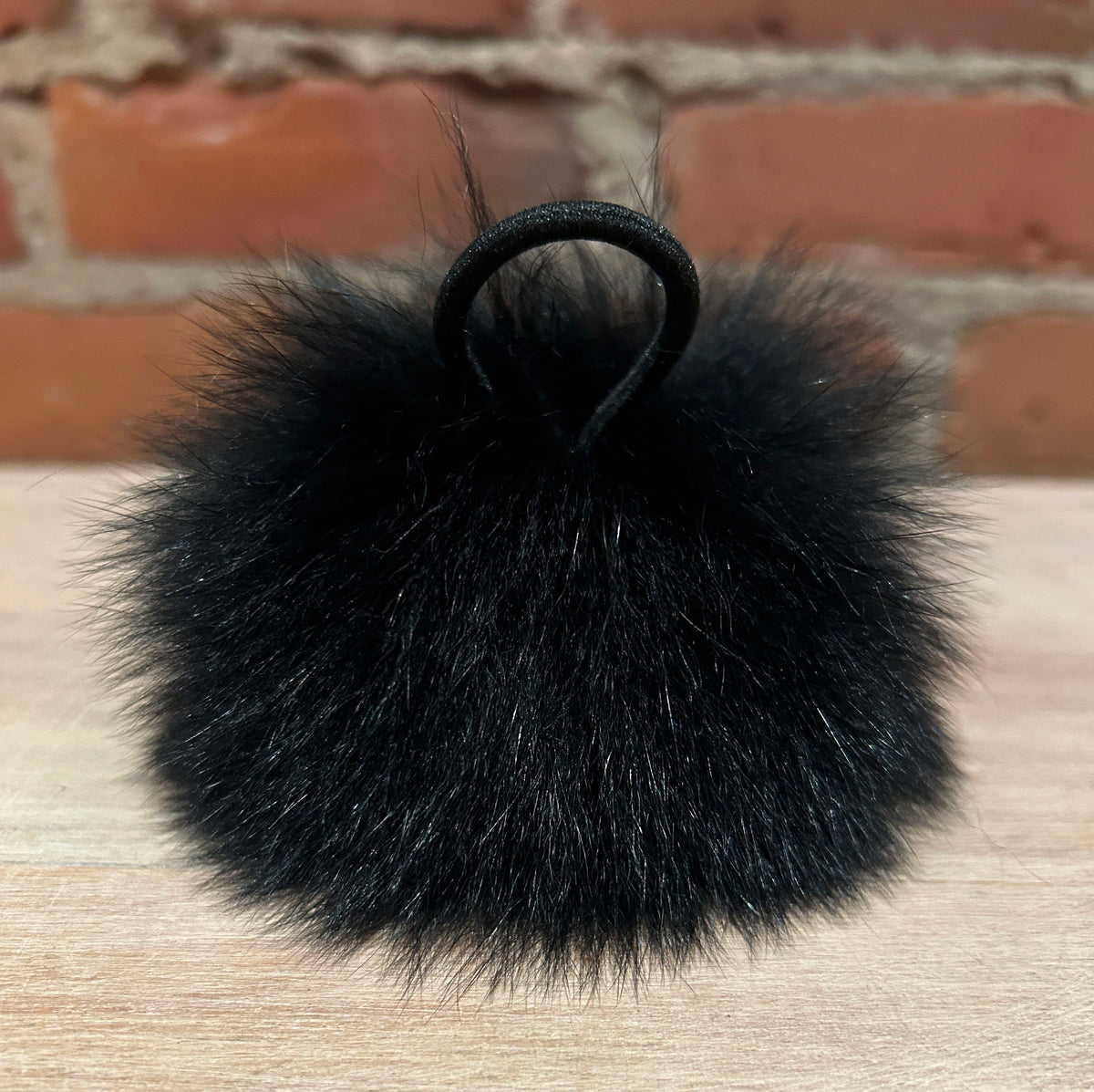 A fluffy black fox fur pom-pom keychain or bag charm with a black elastic hair tie loop, resting on a light wood surface with a blurred brick wall background.