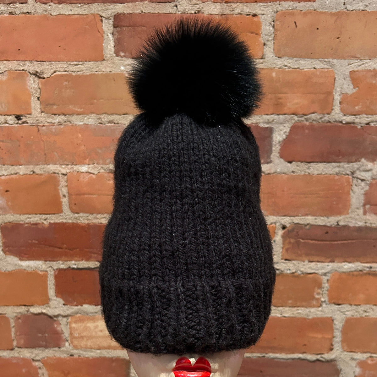 A black ribbed knit beanie with a large black fox-fur pom-pom, displayed on a mannequin head with red lipstick in front of a red brick wall.