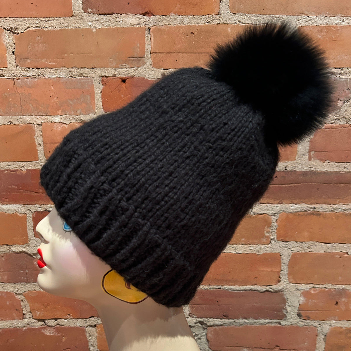 A black ribbed knit beanie with a large black fox fur pom-pom, displayed on a mannequin head against a red brick wall background.