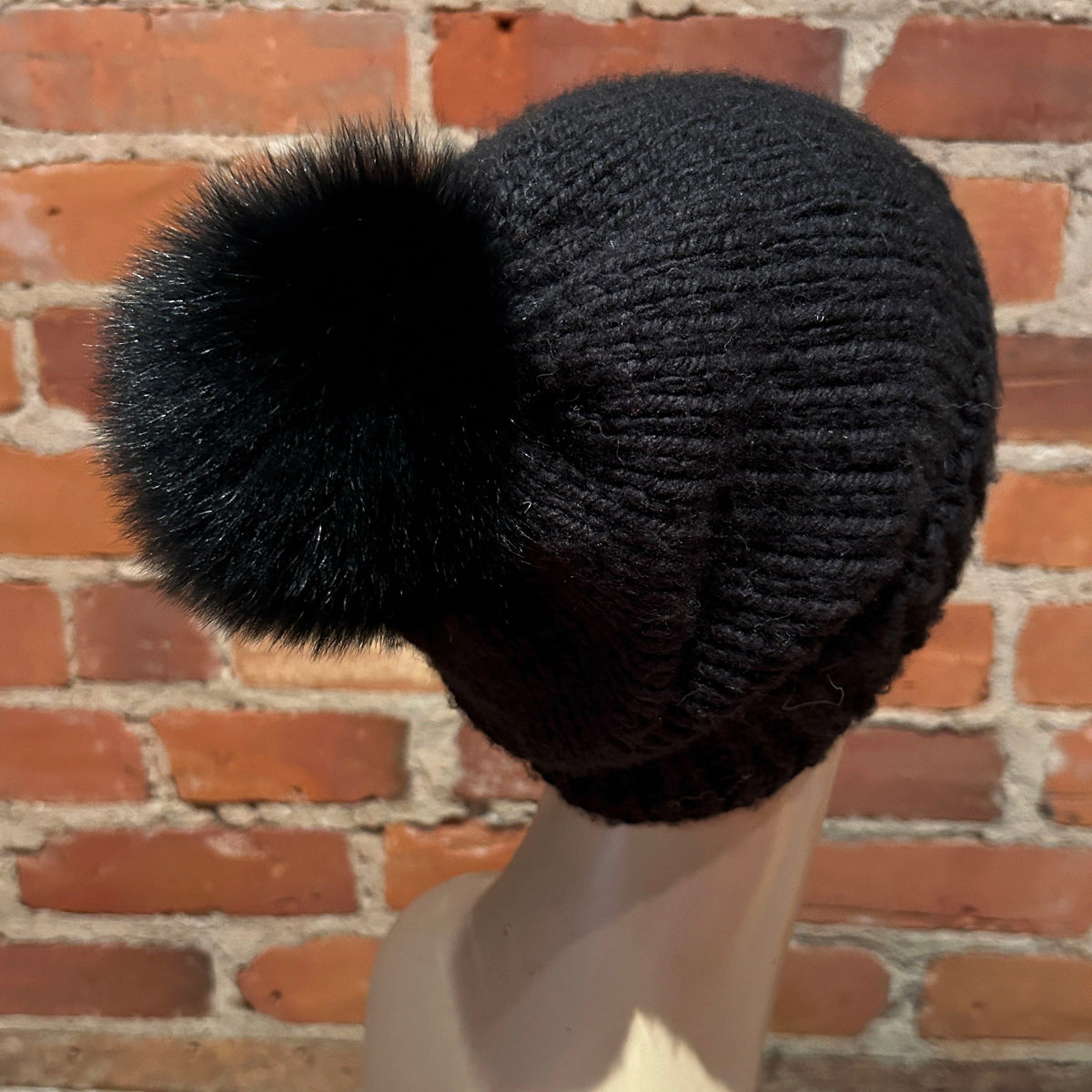 A black ribbed knit beanie with a large fluffy pom-pom, displayed on a mannequin head against a red brick wall background.