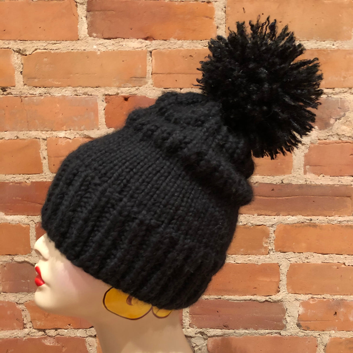 A black ribbed knit pom-pom beanie hat displayed on a mannequin head, set against a red brick wall background.