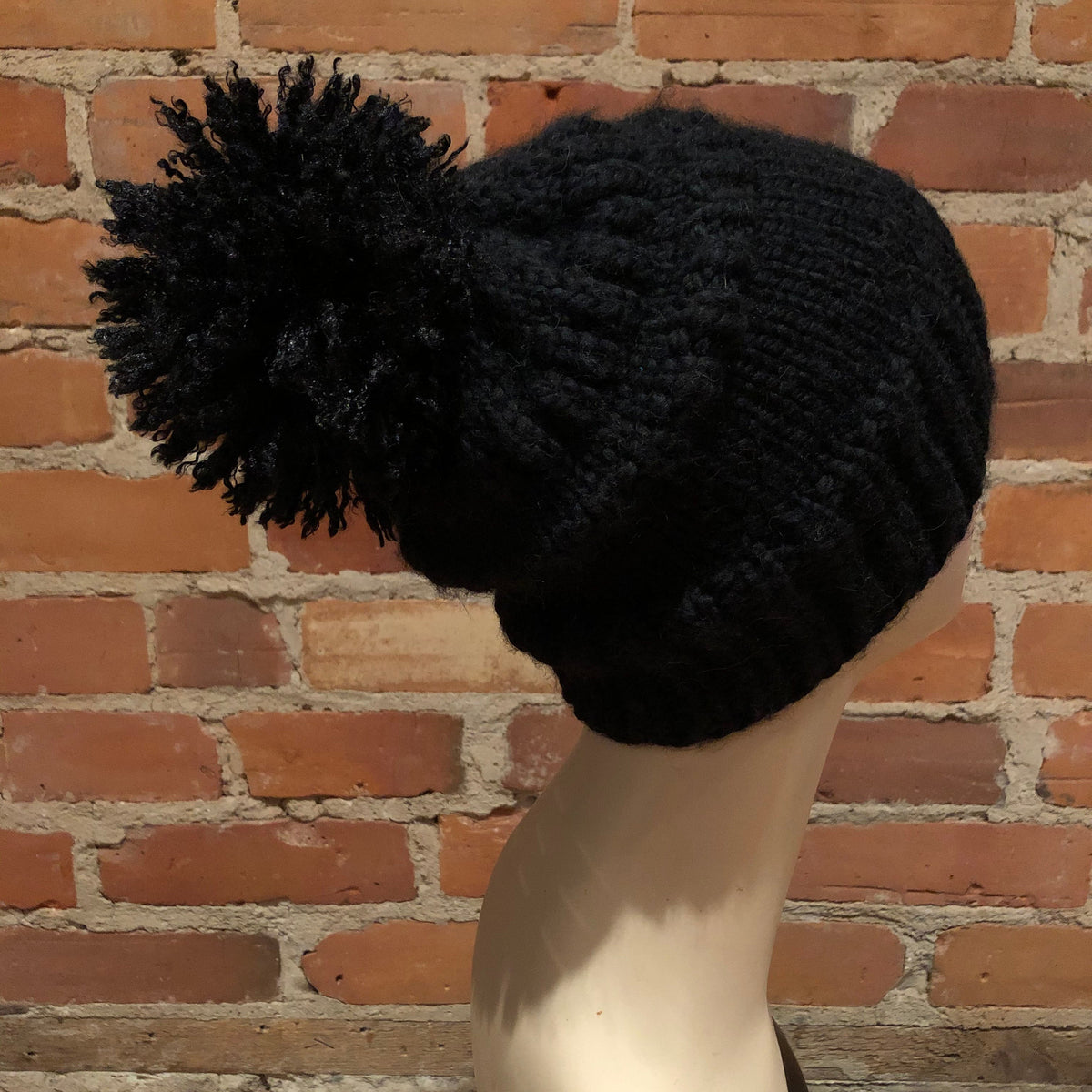 A black chunky knit beanie with a large matching pom-pom, displayed on a mannequin head against a red brick wall background.