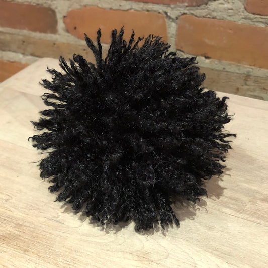 A black curly pom-pom decoration or accessory resting on a light wood surface with a brick wall background.