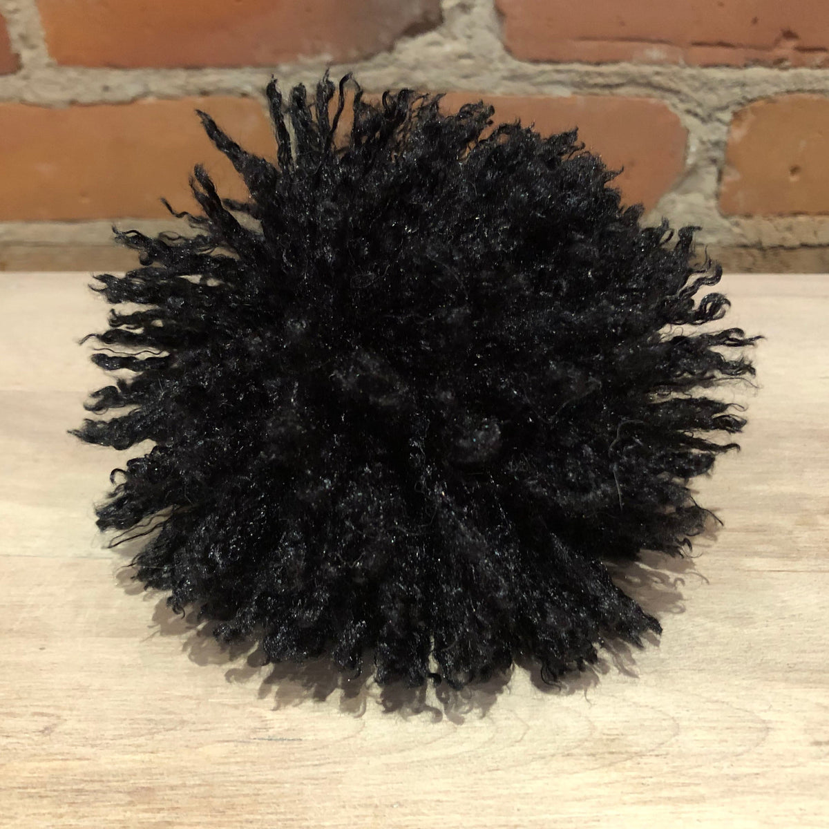 A black curly pom-pom hair accessory with a small black bead at the center, resting on a light wood surface in front of a brick wall background.