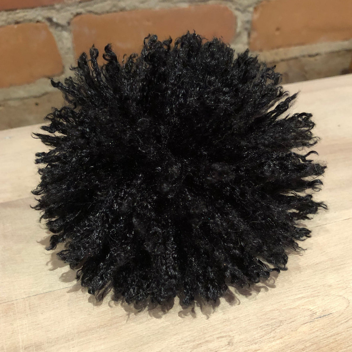 A black curly hair pompadour or hairpiece accessory displayed on a light wood surface with a brick wall background.