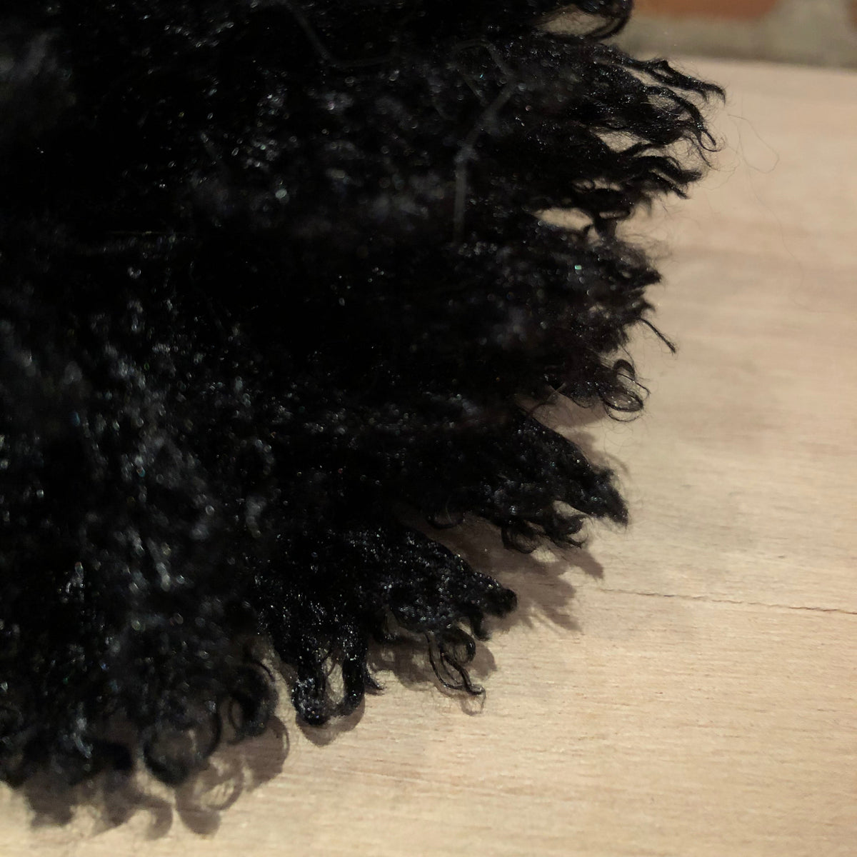 A close-up of a black curly wig or hairpiece resting on a light wood surface with a blurred brick wall in the background.