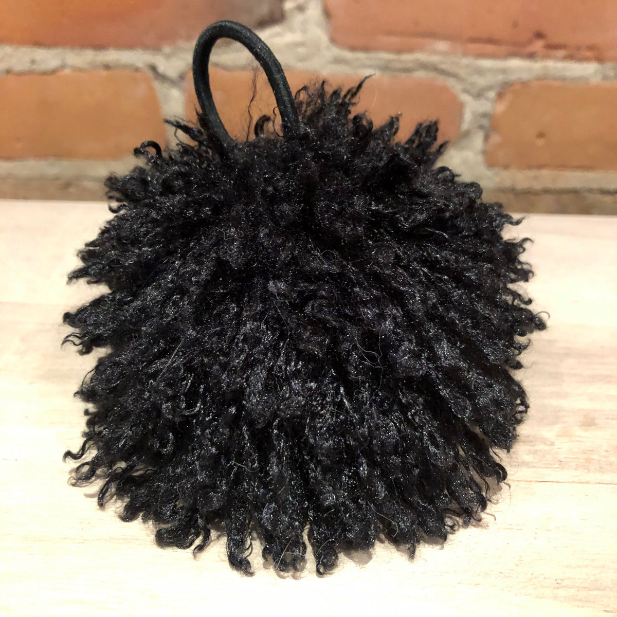 A black curly faux-hair pom-pom accessory with a small black loop handle, displayed on a light wood surface with a brick wall background.