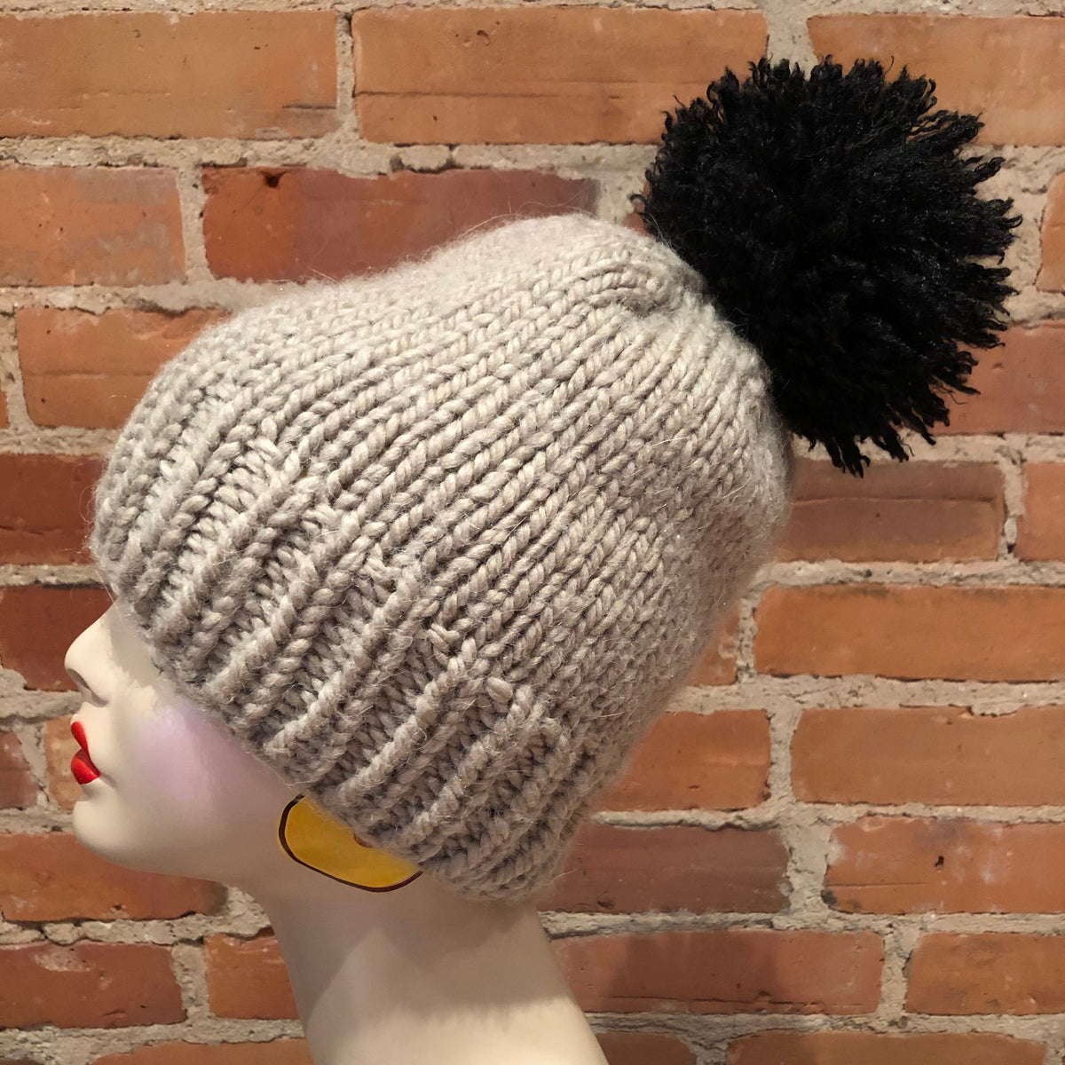 A light gray ribbed knit beanie with a large black pom-pom, displayed on a mannequin head against a red brick wall background.