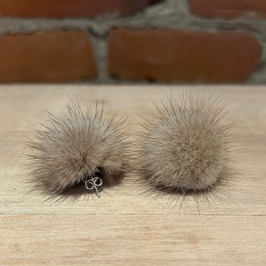 A pair of natural beige mink ear stud earrings with silver posts, displayed on a light wooden surface with a blurred brick wall background.
