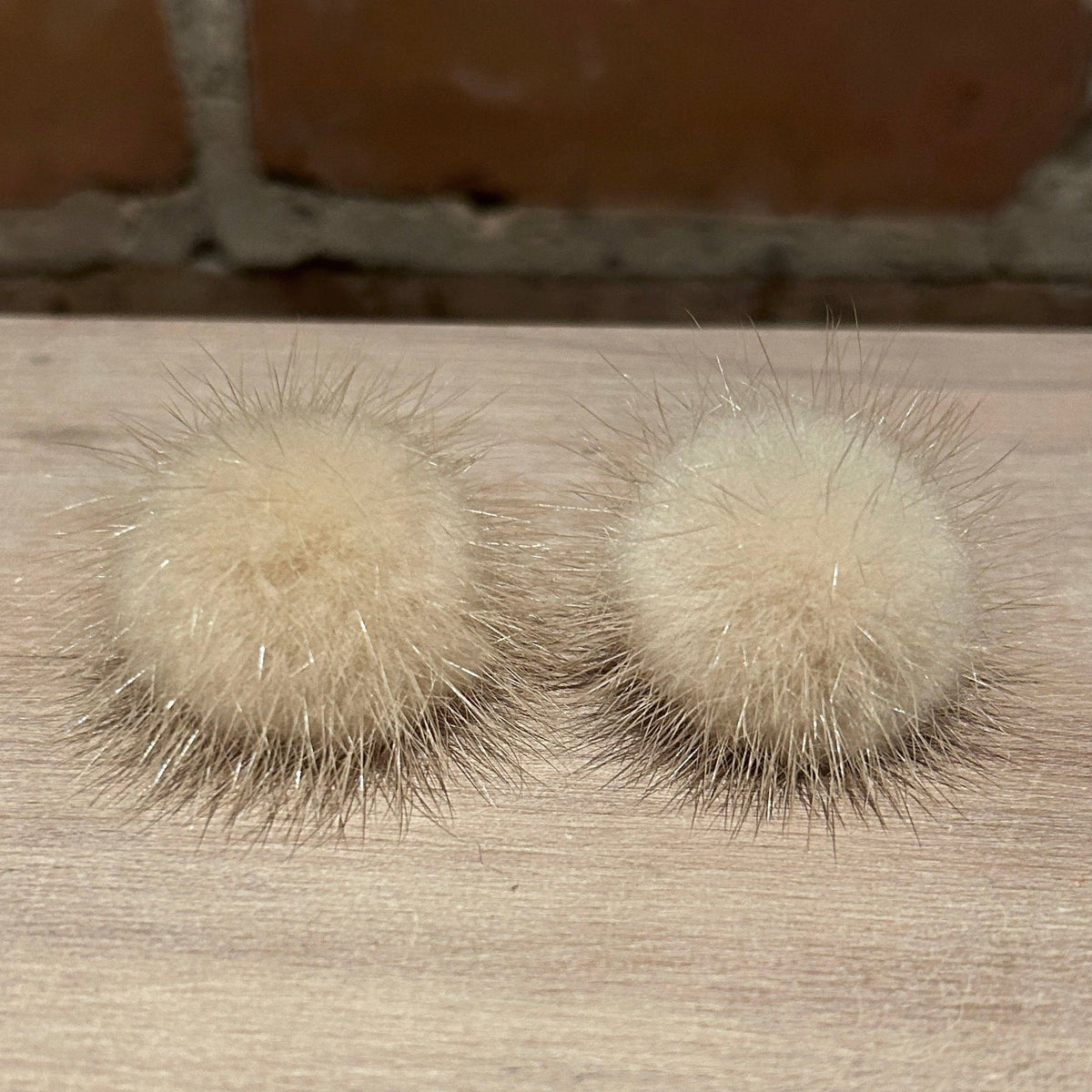 A pair of fluffy pom-pom stud earrings made from cream-colored fur, displayed on a light wood surface with a blurred brick wall in the background.