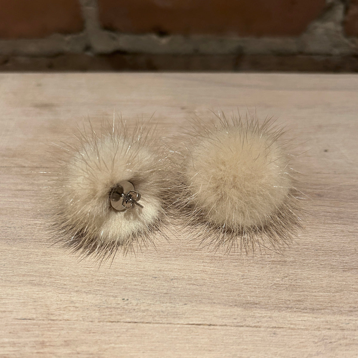 A pair of fluffy pom-pom earrings in a light cream or beige mink fur material, with a small silver metal ring detail on one earring, resting on a light wood surface.