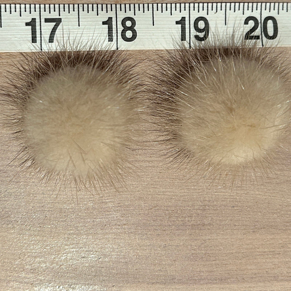 Two round beige mink fur pom-pom earrings s displayed on a light wood surface with a measuring ruler at the top showing centimeter markings.