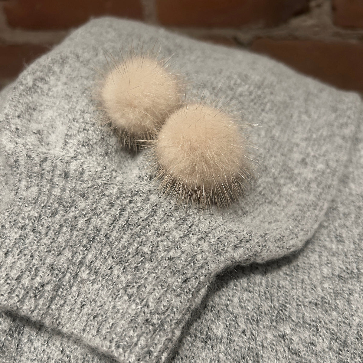 A close-up of a gray knit garment, likely a sweater or hat, featuring two fluffy cream mink pom-pom earrings on the front, set against a rustic brick wall background.