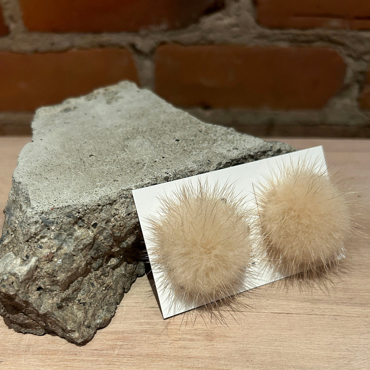A pair of fluffy pom-pom stud earrings in a light beige/tan recycled mink fur,, displayed on a small white card. The card is placed on a light wood surface next to a large gray stone, with a blurred brick wall in the background.