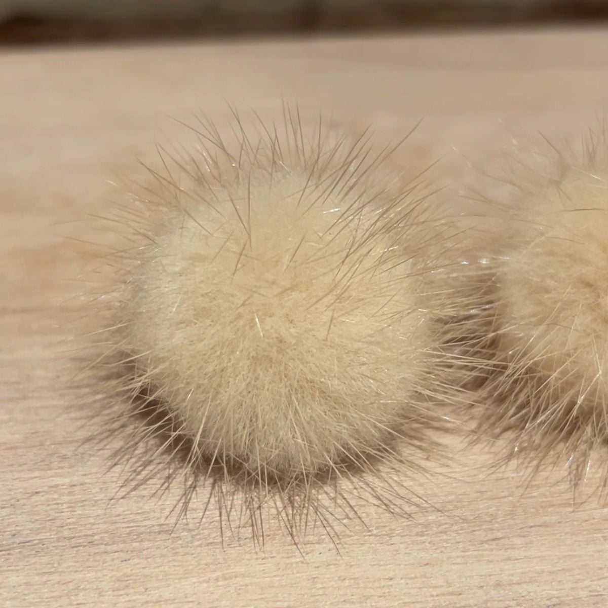 A close-up of a fluffy pom-pom accessory in a light cream or beige color, resting on a light wood surface with a blurred brick wall in the background.