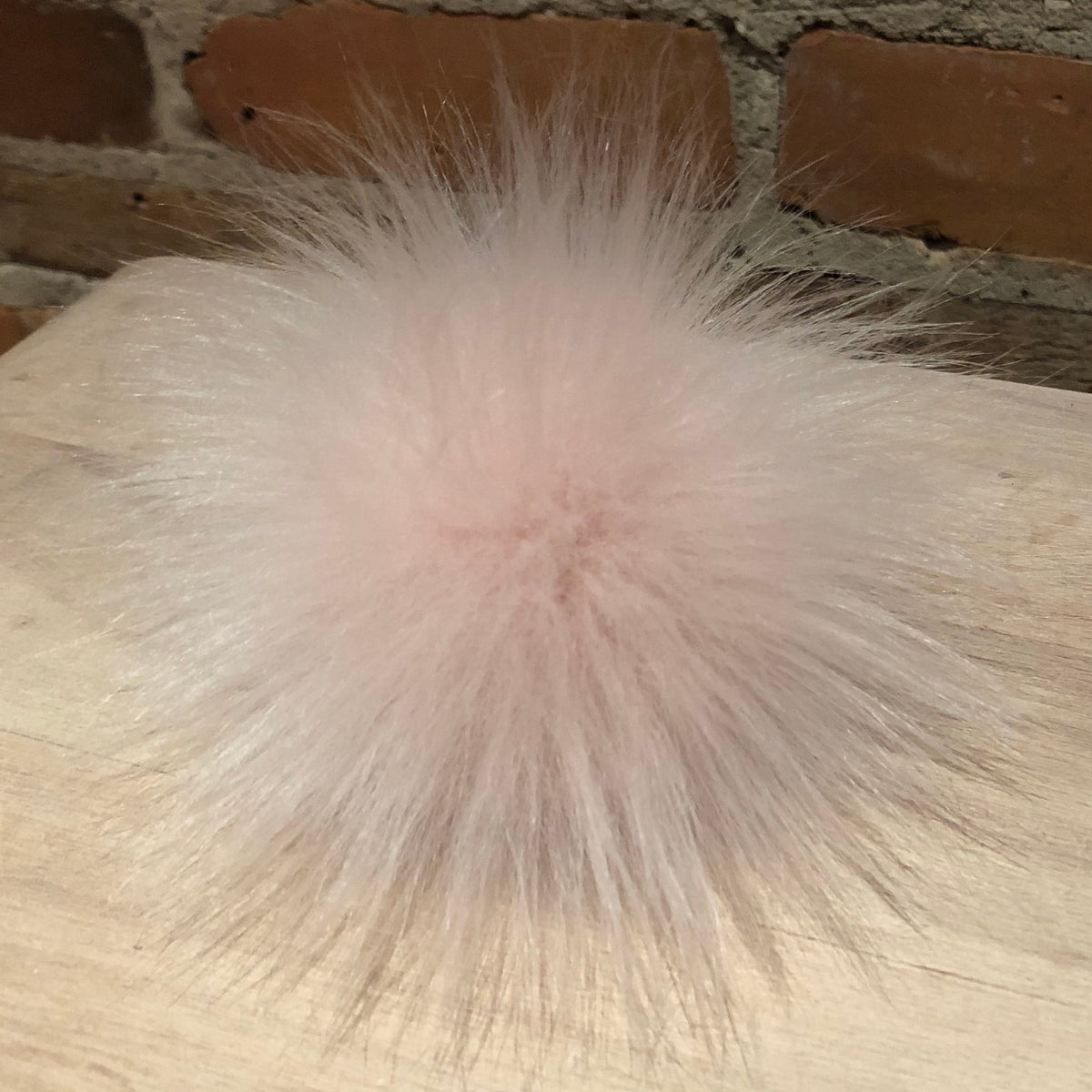 A fluffy, pale pink faux fur pom-pom accessory resting on a light wood tabletop with a brick wall background.