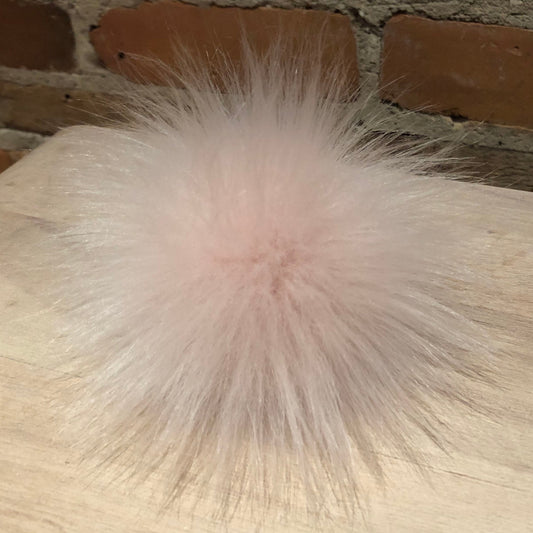 A fluffy, pale pink faux fur pom-pom accessory resting on a light wood tabletop with a brick wall background.