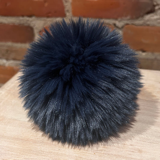 A dark navy blue fluffy pom-pom accessory resting on a light wood surface with a brick wall background.