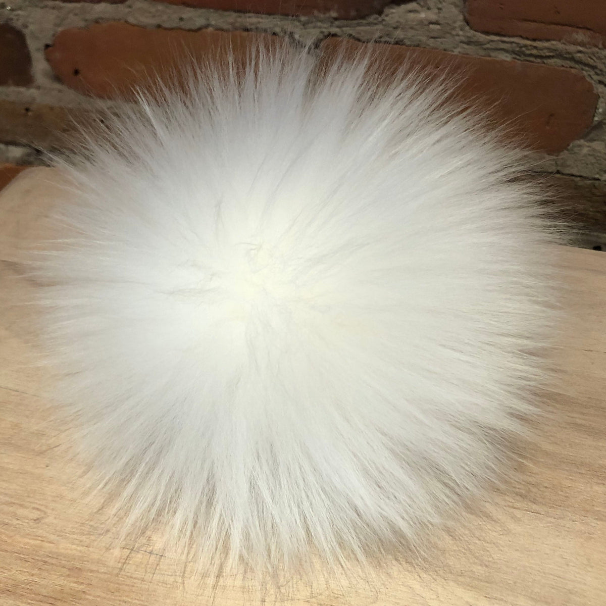 A fluffy white lamb fur pom-pom ball resting on a light wood surface with a brick wall background.