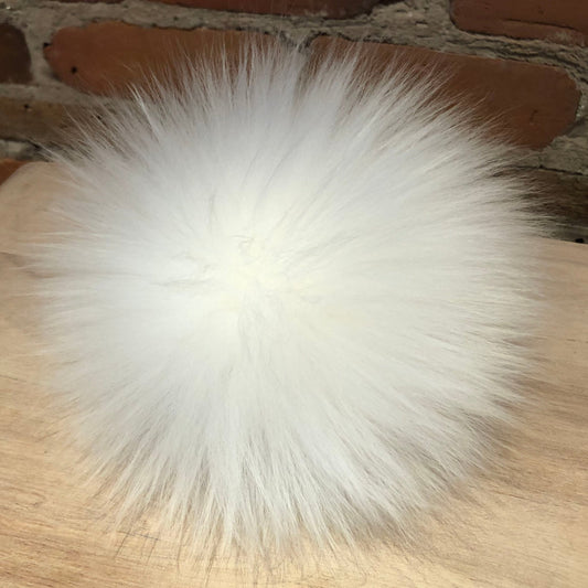 A fluffy white lamb fur pom-pom ball resting on a light wood surface with a brick wall background.