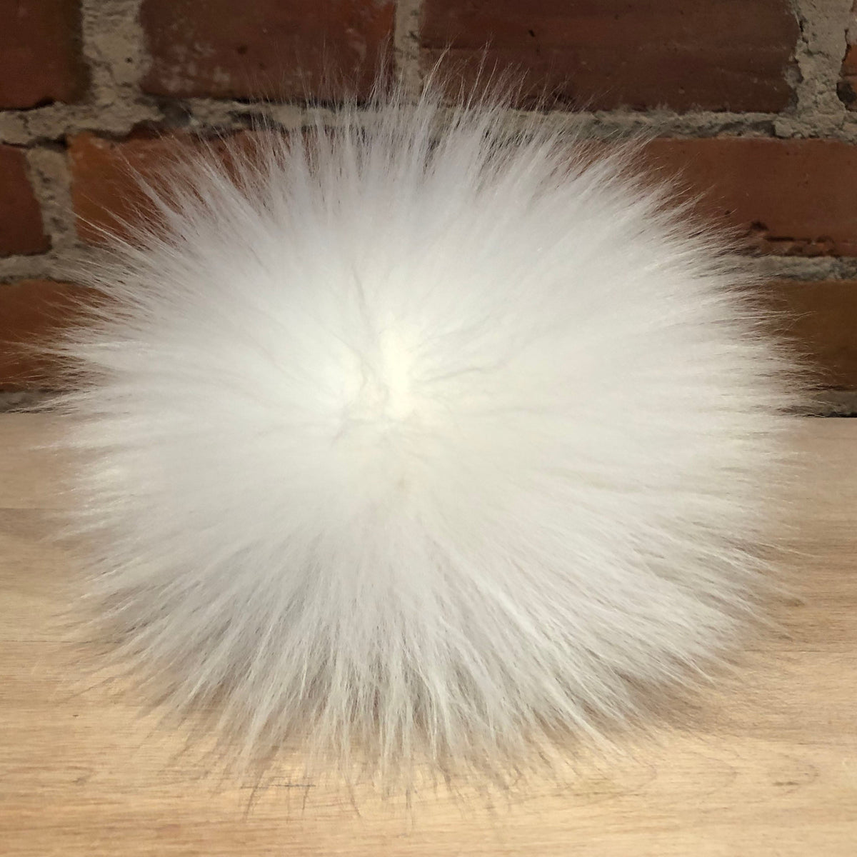 A fluffy white lamb fur pom-pom ball resting on a light wood surface with a red brick wall background.