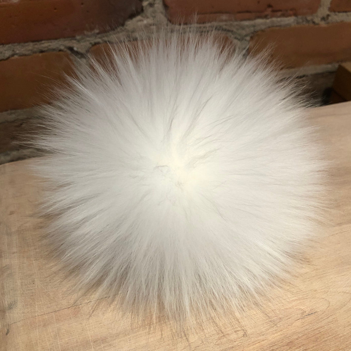 A fluffy white lamb fur pom-pom ball resting on a light wood surface with a brick wall in the background.