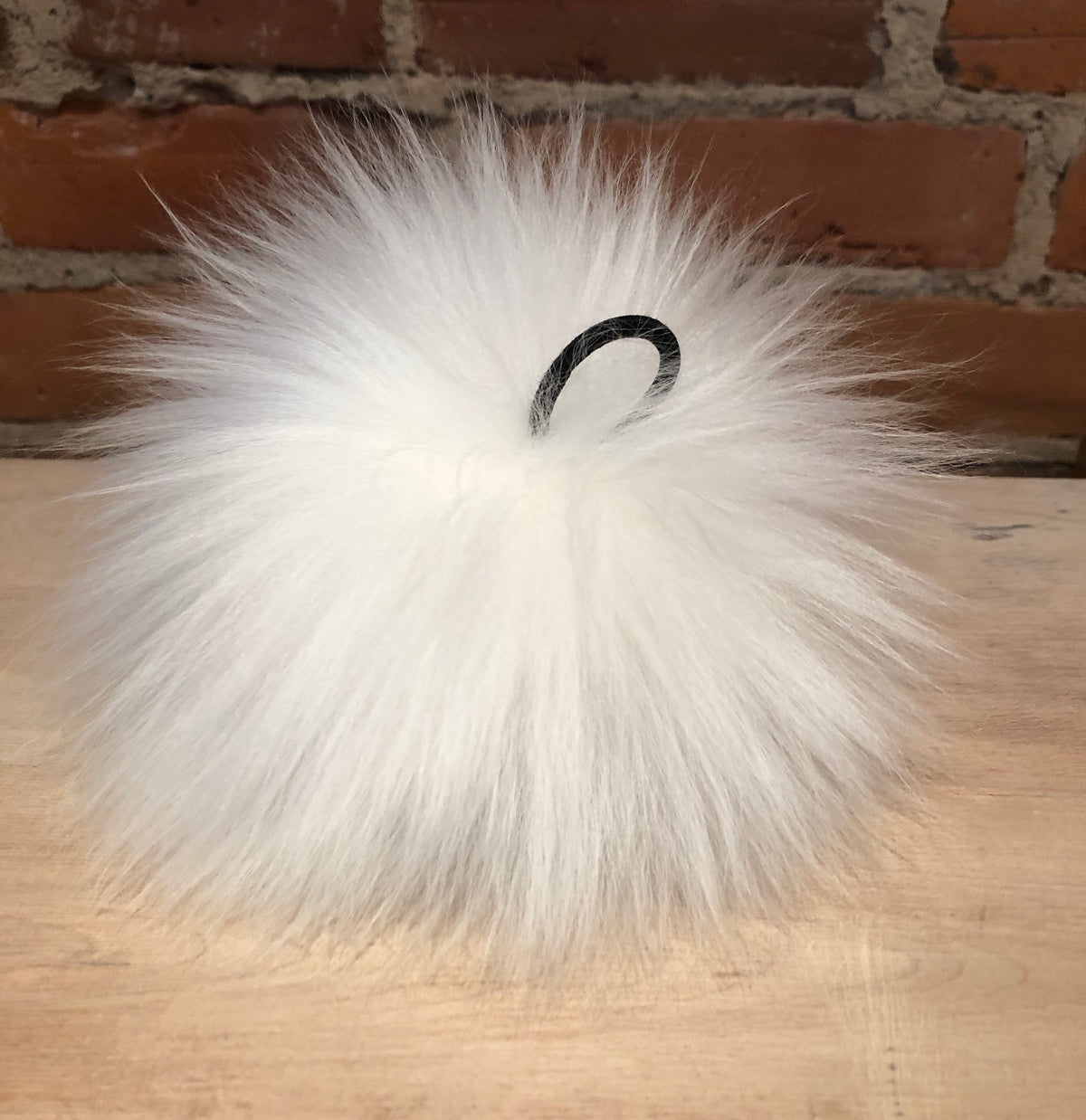 A fluffy white lamb fur pom-pom bag charm with a black elastic loop, resting on a light wood surface with a brick wall background.