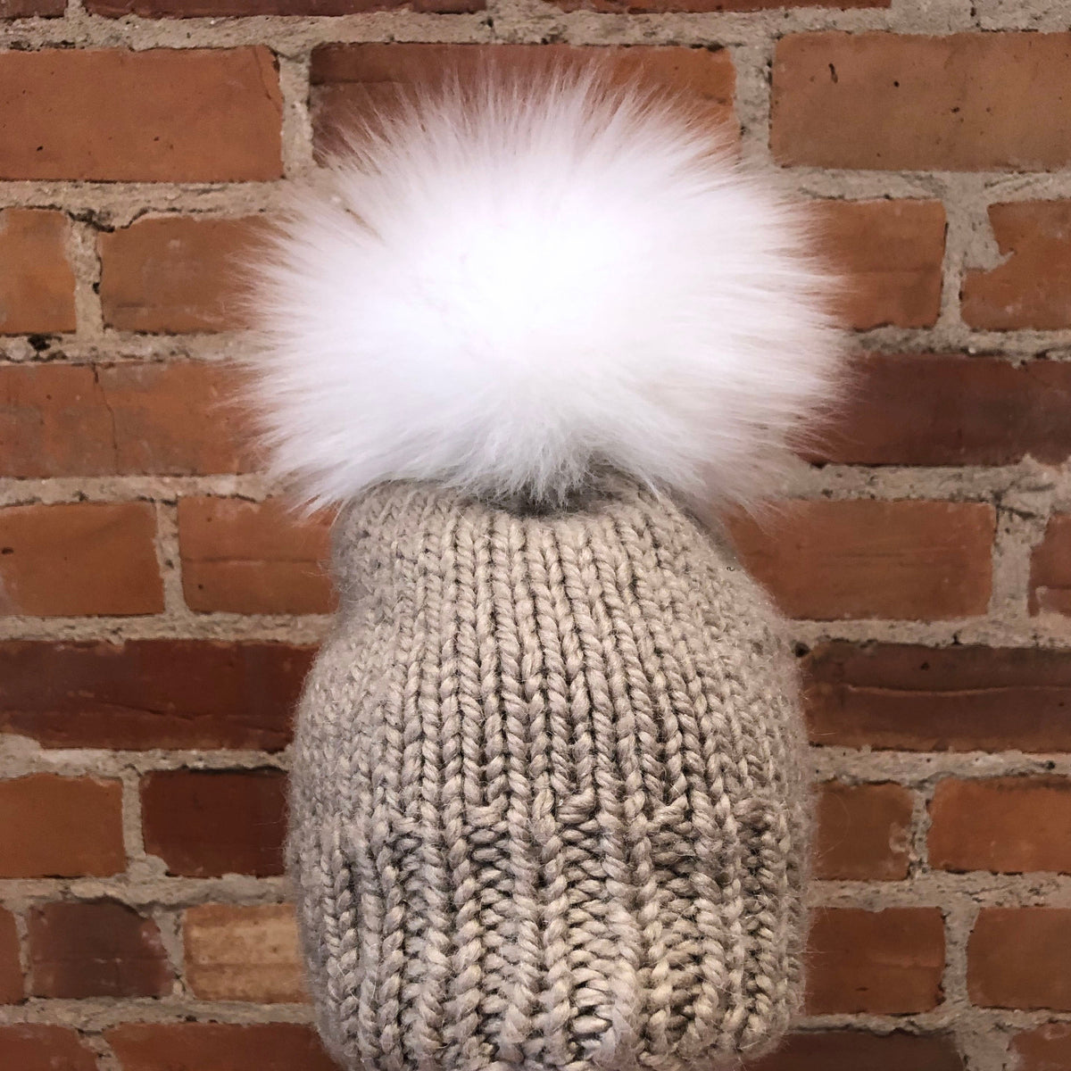 A chunky knit beanie in a light taupe-gray color with a large fluffy white lamb-fur pom-pom, displayed on a mannequin head against a red-brown brick wall background.