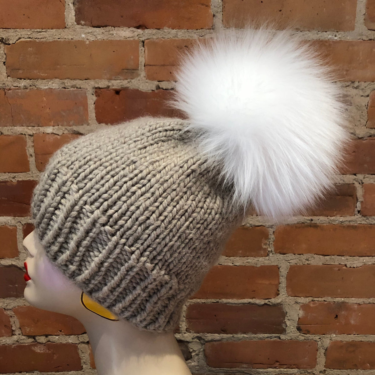 A beige ribbed knit beanie with a large fluffy white lamb-fur pom-pom, displayed on a mannequin head against a red-brown brick wall background.